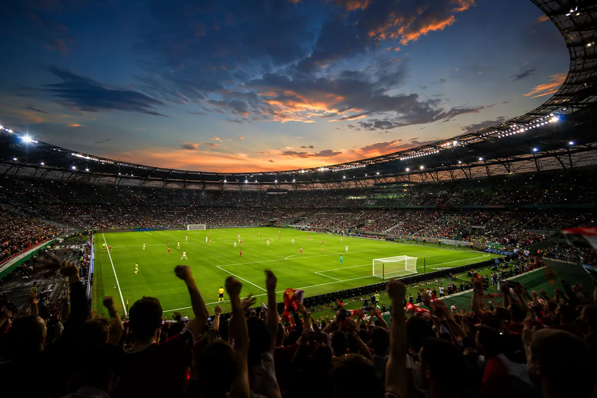 Wide shot of a vibrant football stadium at dusk, bright stadium lights illuminating the gr