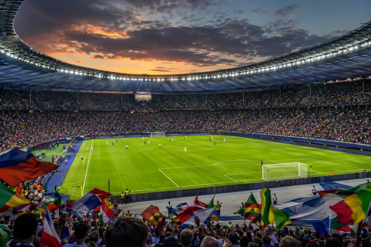 Wide shot of a modern football stadium at dusk, with colorful crowd of fans holding generi