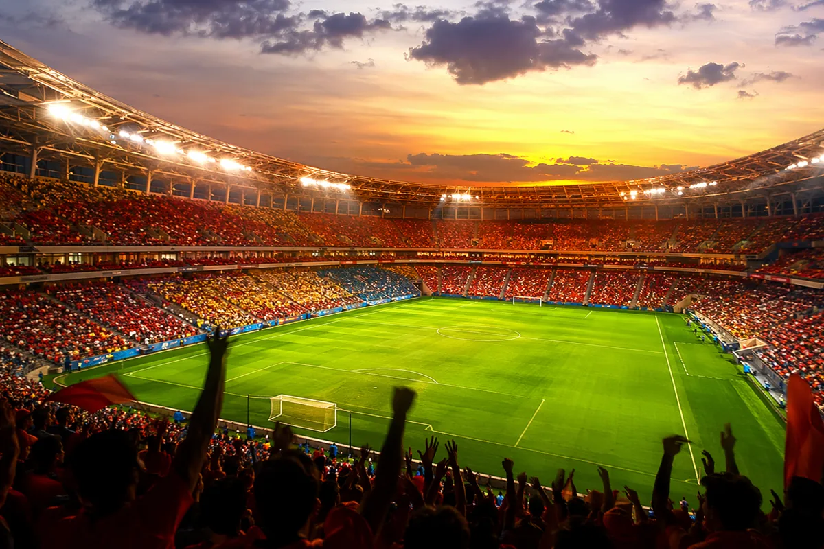 Wide shot of a modern football stadium at dusk, vibrant green pitch, empty seats in bright