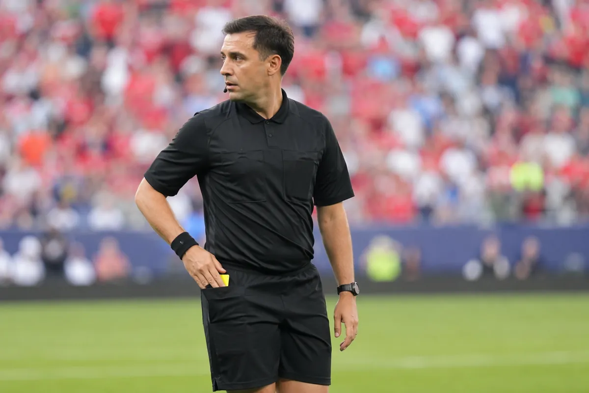 Professional football referee standing on green soccer pitch, hand resting near pocket whe