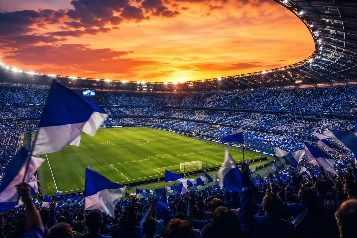 Wide shot of a vibrant football stadium at sunset, cheering fans waving blue and white fla