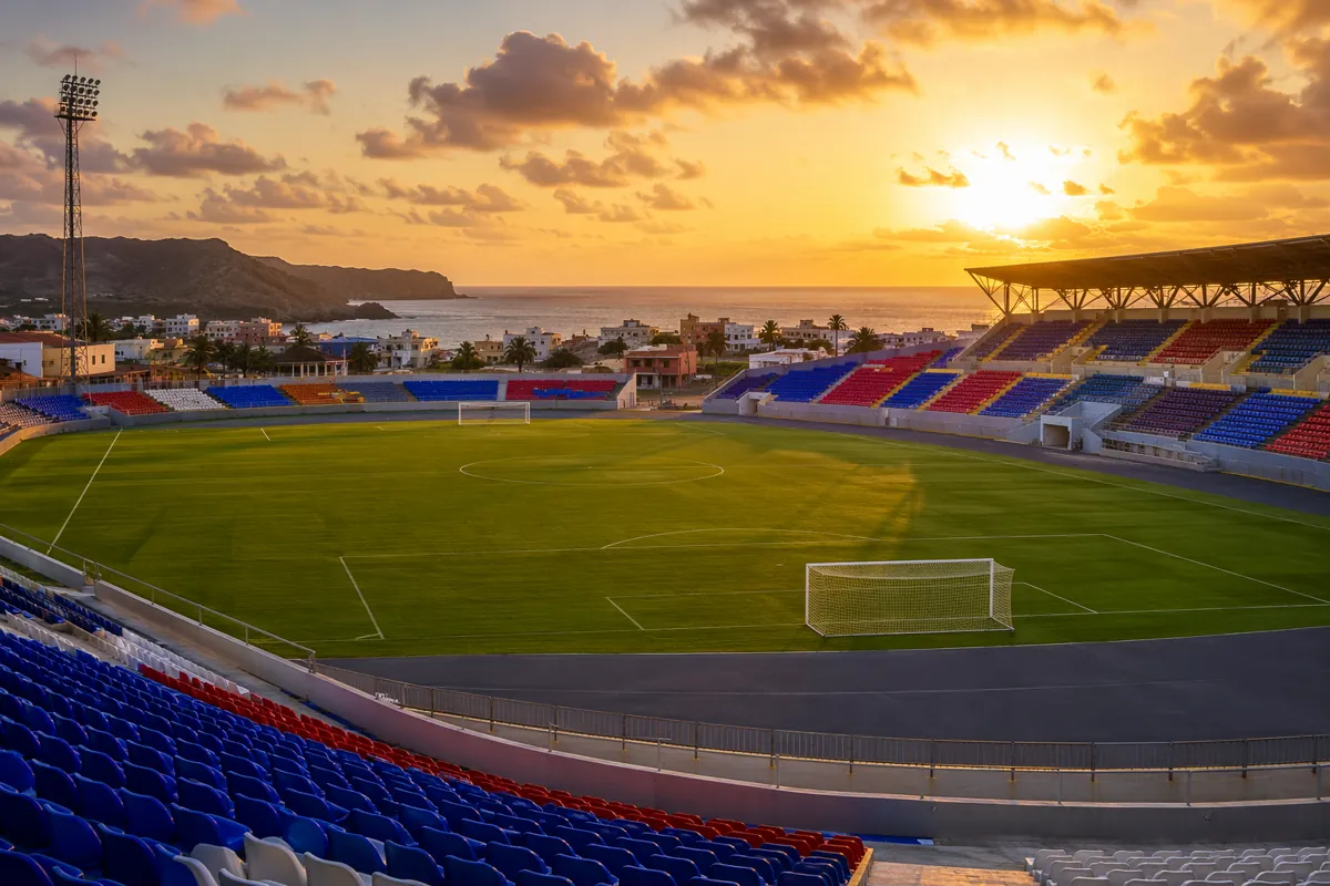 Wide shot of a vibrant coastal football stadium in Cape Verde at sunset, empty green pitch