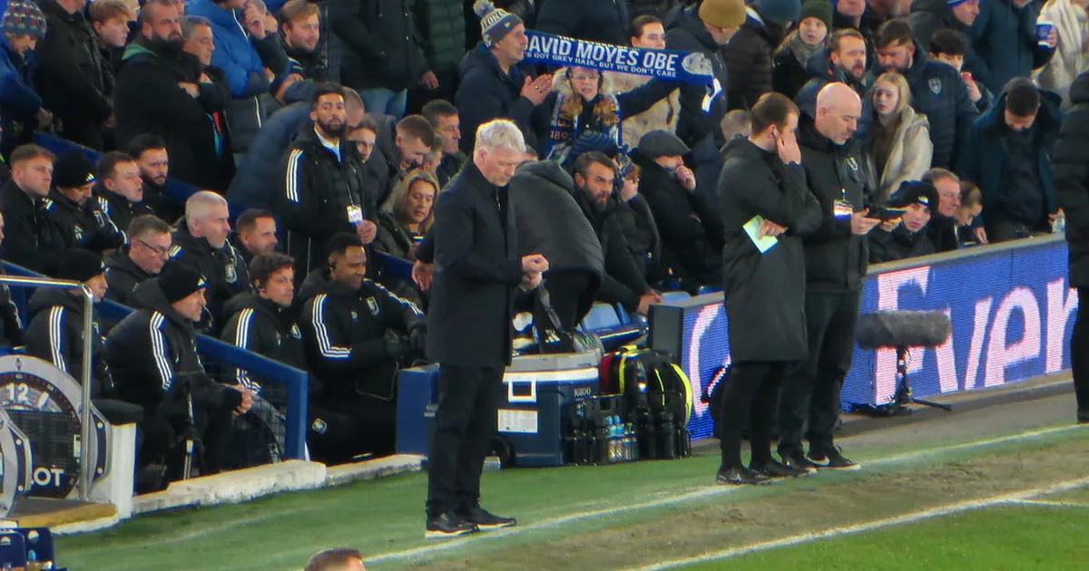 A wide shot of a professional football manager standing in the dugout of a modern football