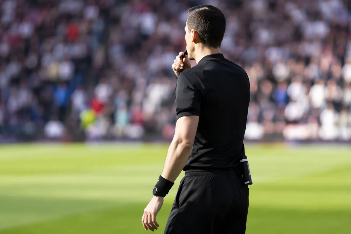 Professional football referee wearing black uniform holding a whistle, standing on a green