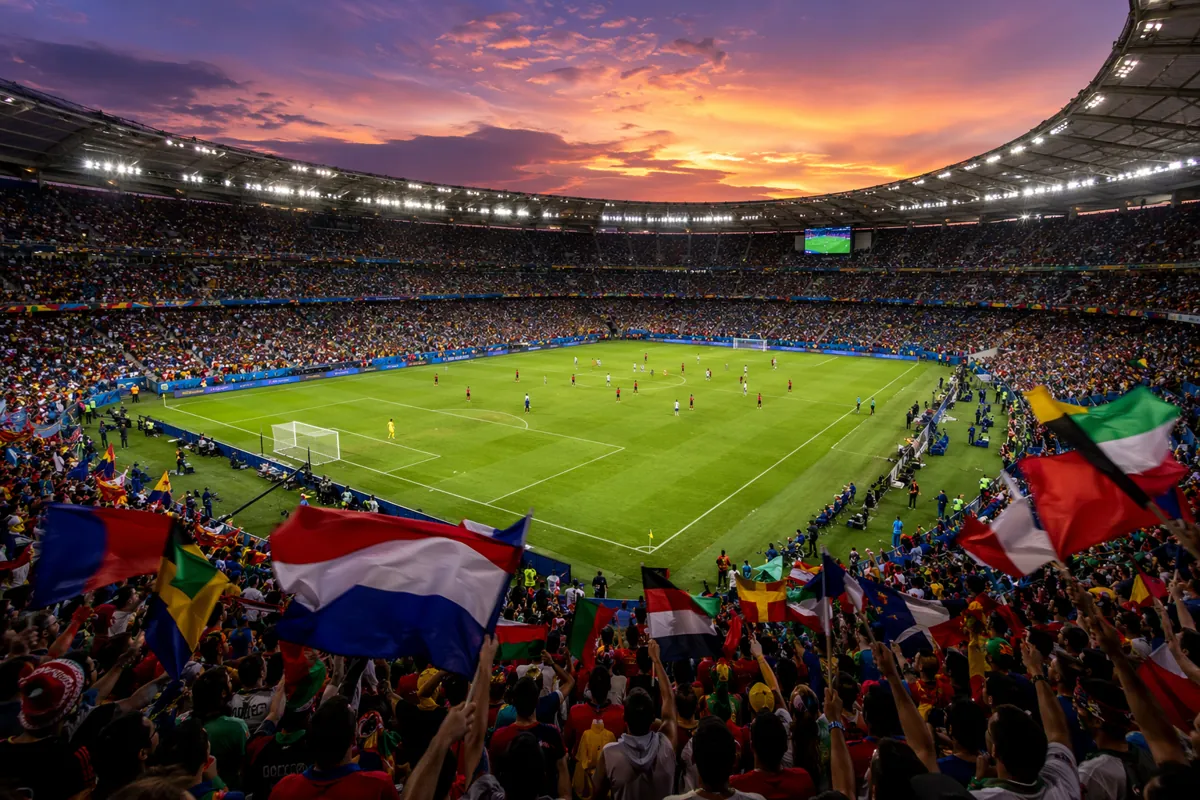 Wide shot of a vibrant World Cup football stadium at dusk, with colorful fan flags waving