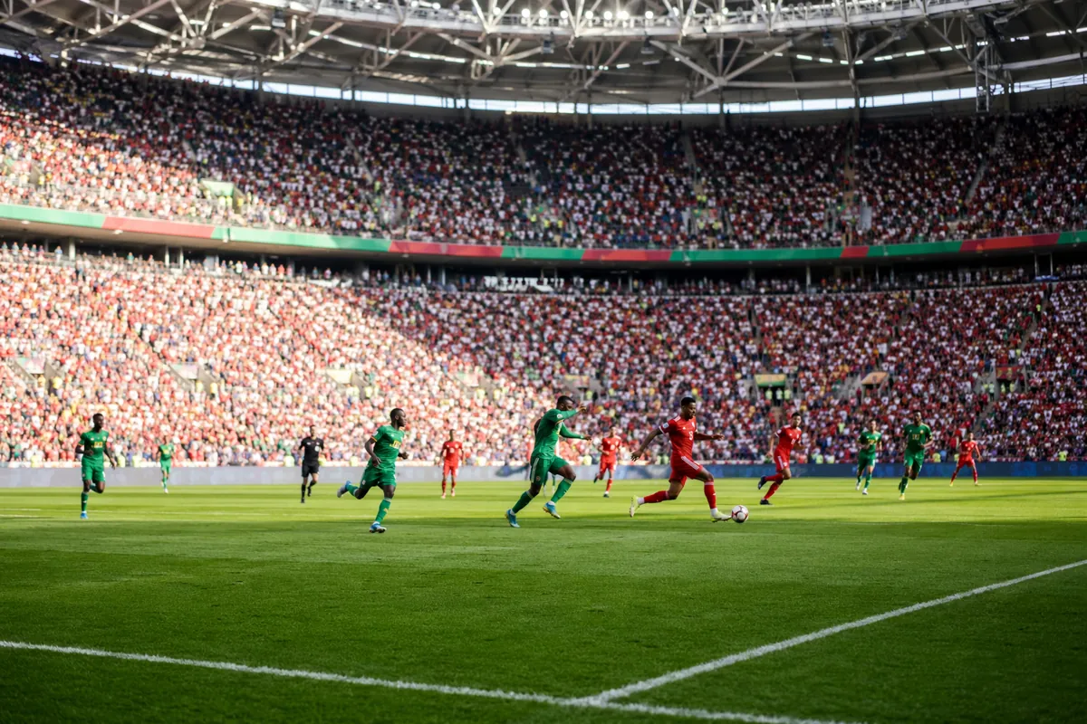 Wide shot of professional football match in a sunlit modern stadium, players in red and gr
