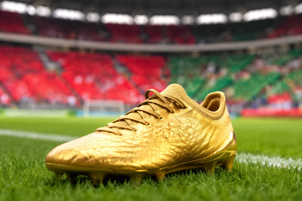 Close-up of a golden football boot placed on well-maintained green grass pitch, blurred ba