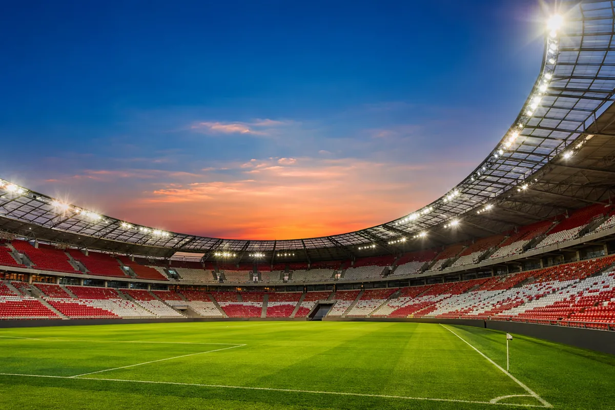 Wide shot of a vibrant football stadium at dusk, with green pitch, spectator seats in red
