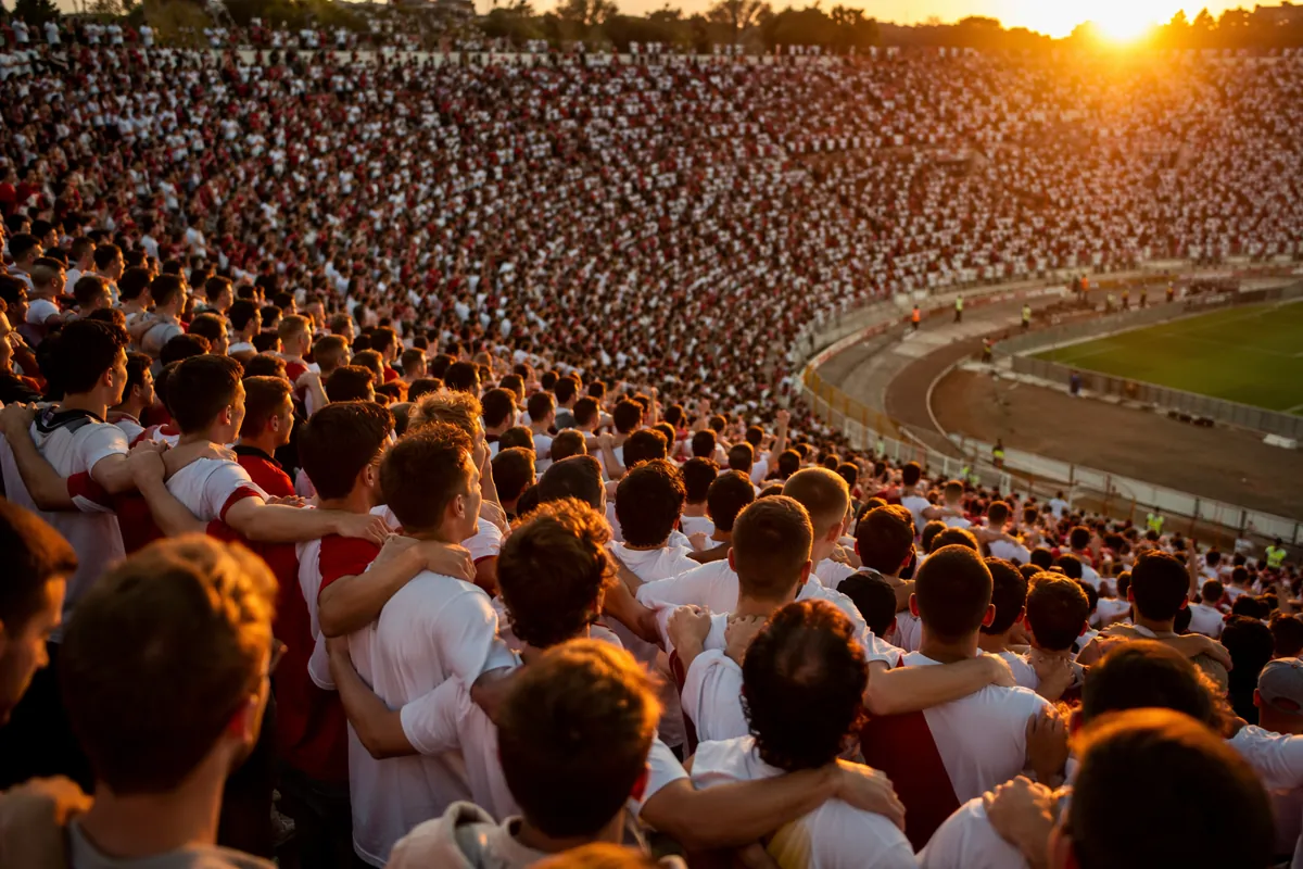 Wide shot of a packed football stadium crowd during golden hour, thousands of fans wearing