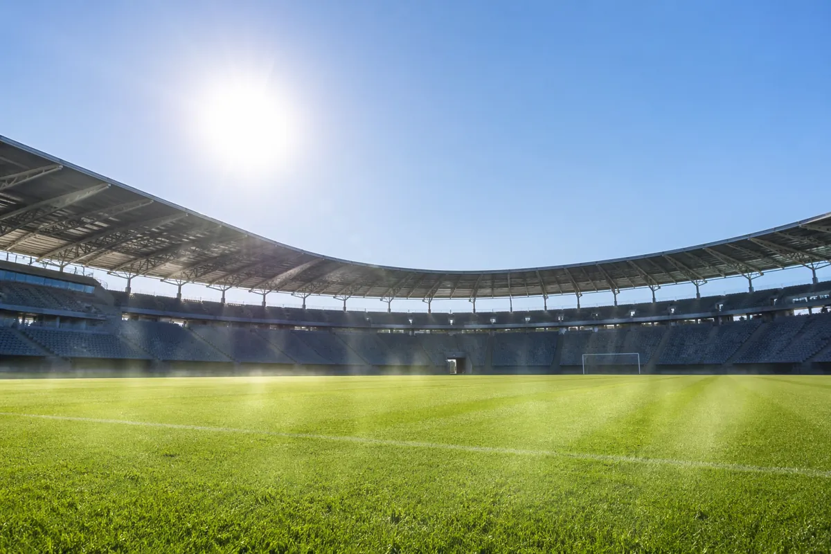 Wide shot of a modern open-air football stadium under bright scorching sun, clear blue sky