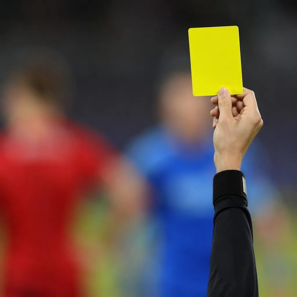 Close up shot of a soccer referee's hand showing a yellow card, blurred players