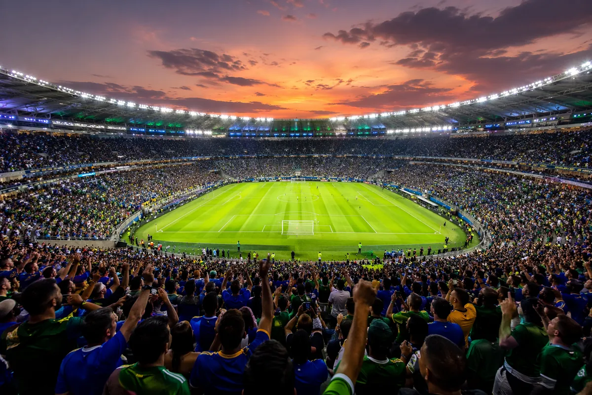 Wide shot of a vibrant World Cup football stadium at dusk, packed with cheering fans weari