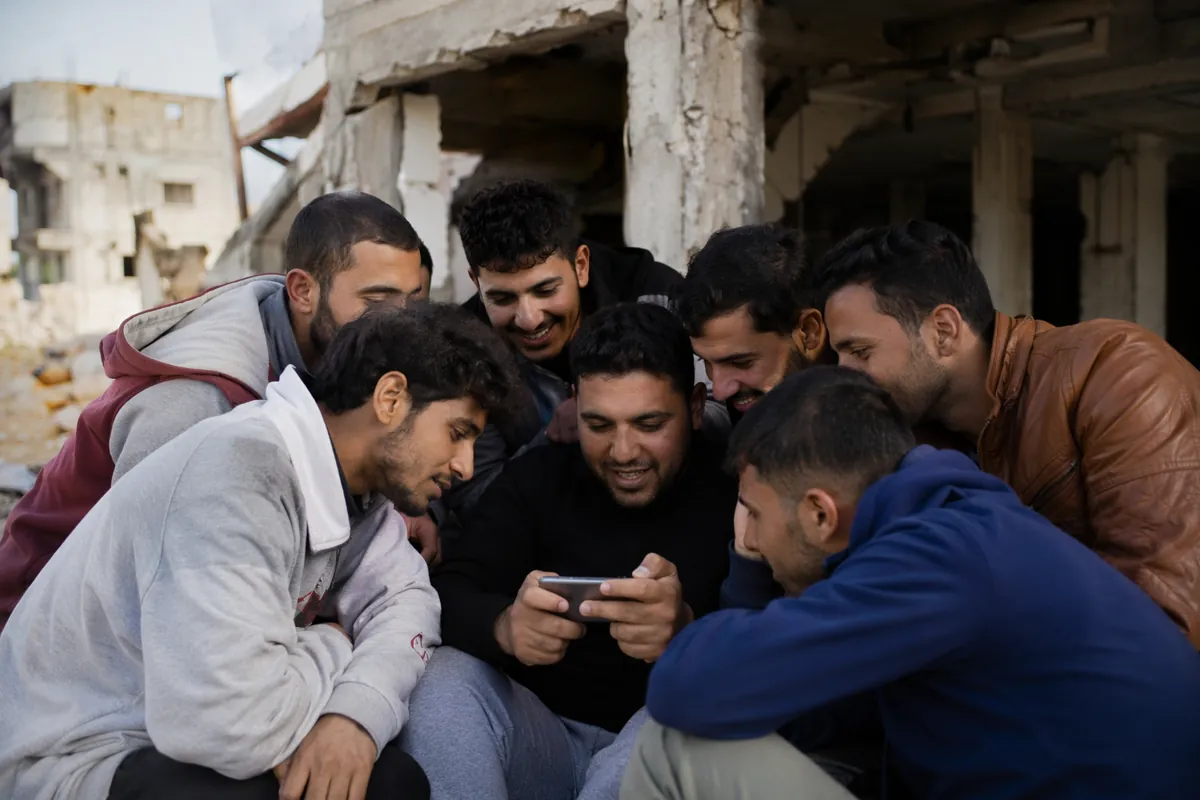 A group of young Arab men huddled together around a small mobile phone in a partially dama