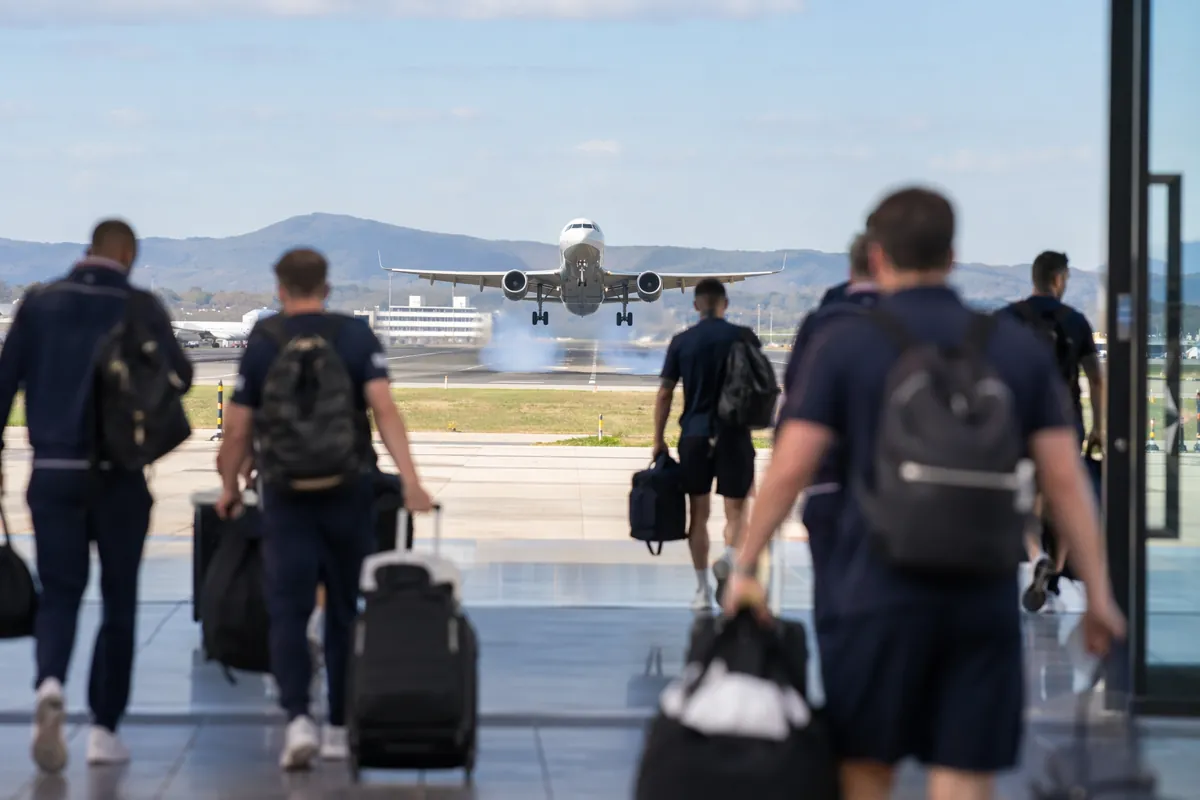 Wide shot of a commercial airplane landing on a sunny airport runway, with blurred footbal