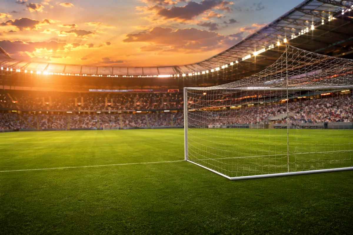 Professional football stadium at dusk with warm golden light, green grass pitch, empty goa