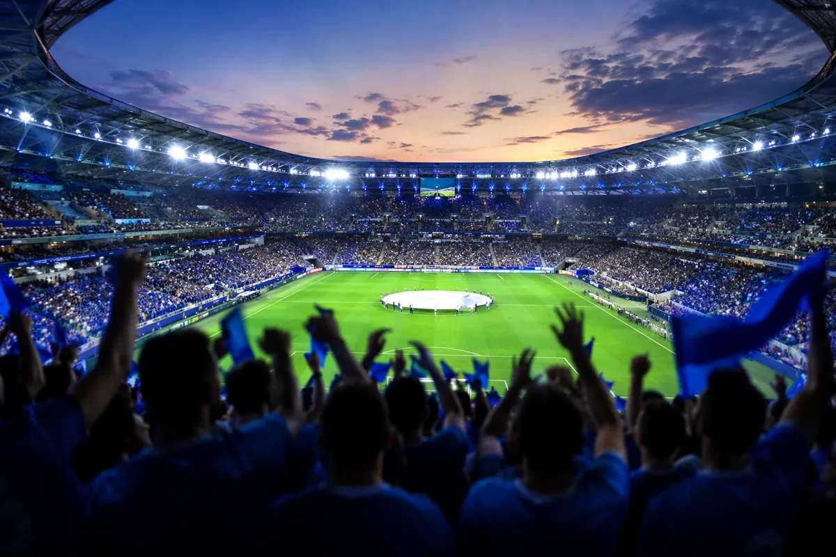 Wide shot of a modern football stadium at dusk, bright stadium lights on, blurred silhouet