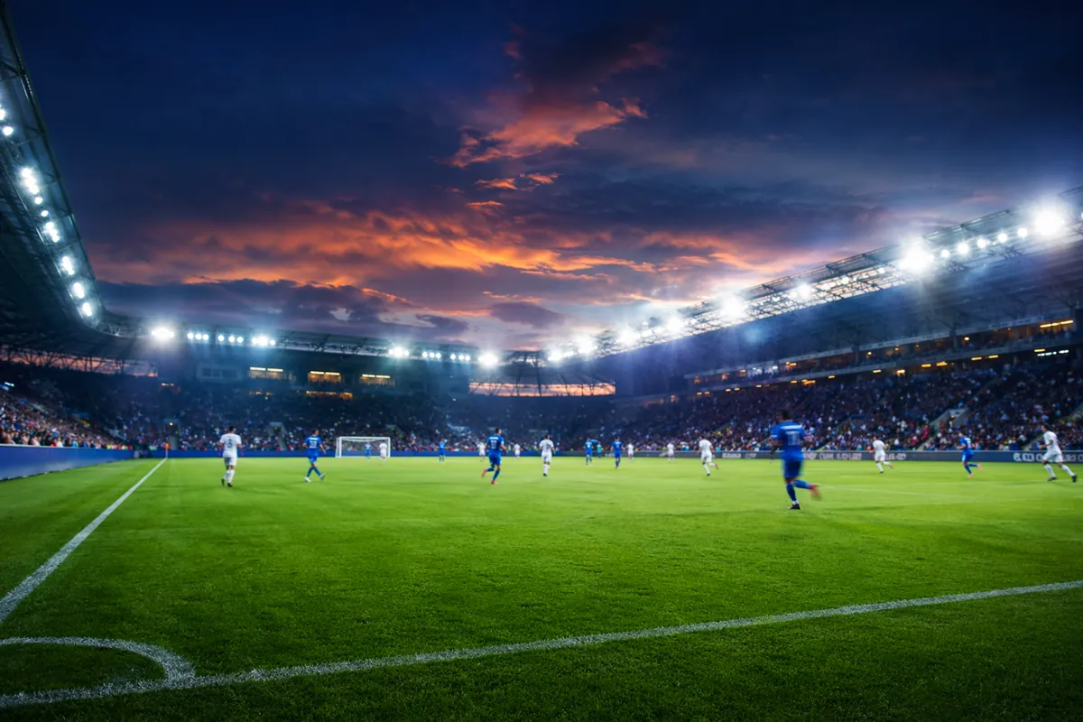 Wide shot of a professional football stadium at dusk, bright floodlights illuminating the
