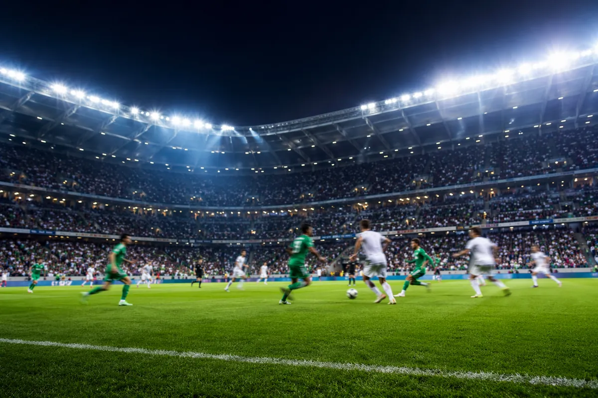 Wide shot of a professional football stadium during a friendly match, blurred players in g