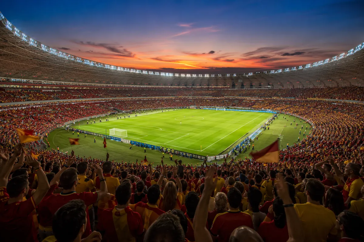 Wide shot of a professional football stadium at dusk, filled with cheering fans wearing re