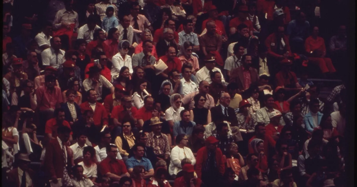 Wide shot of a packed football stadium during sunset, red team fans waving flags and cheer