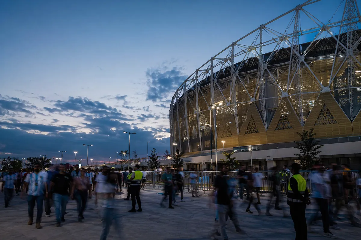 Dusk view of a modern World Cup stadium outer area with blurred fans walking, security gua