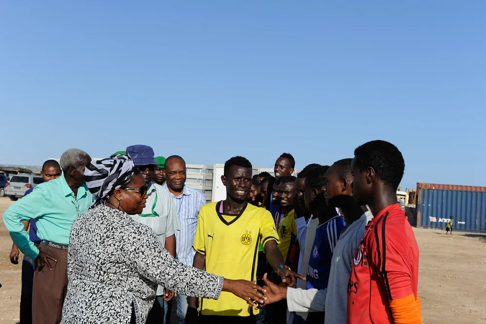 Wide shot of a youth football match in progress, stands with sparse casual spect