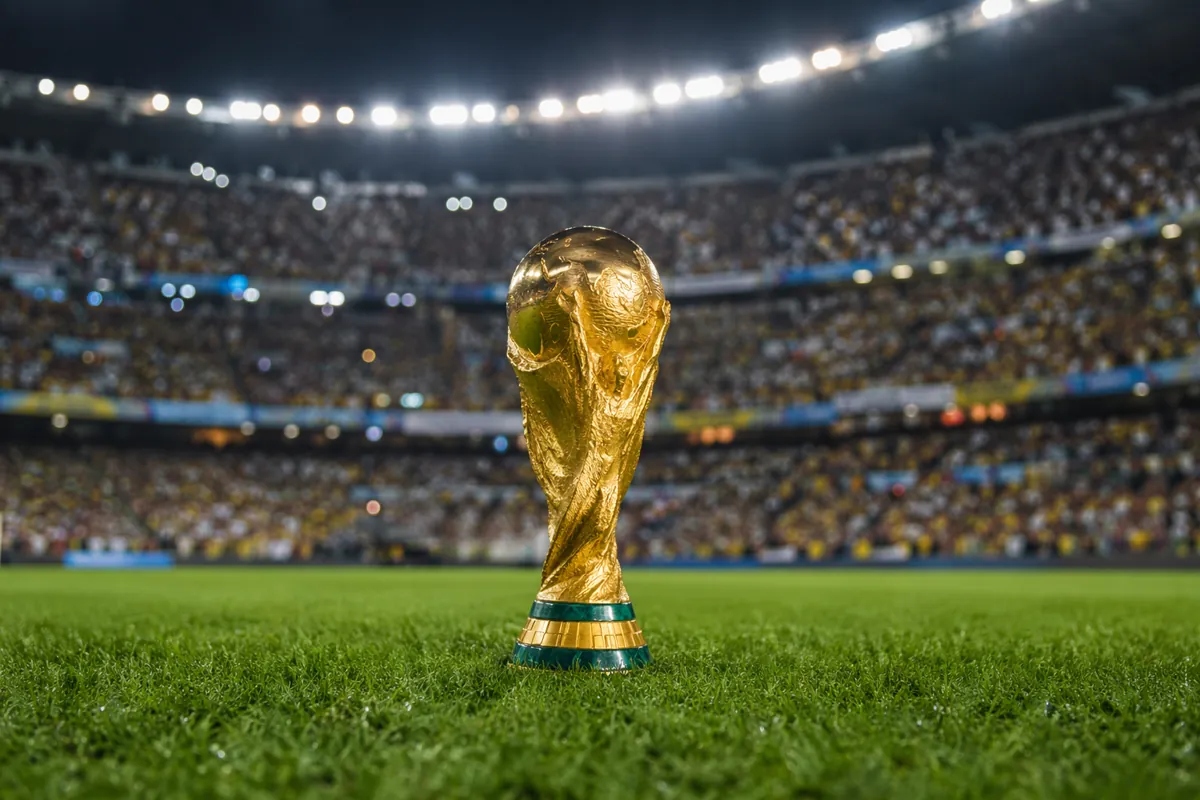 Wide shot of a golden world cup trophy placed on a green football field, with blurred chee