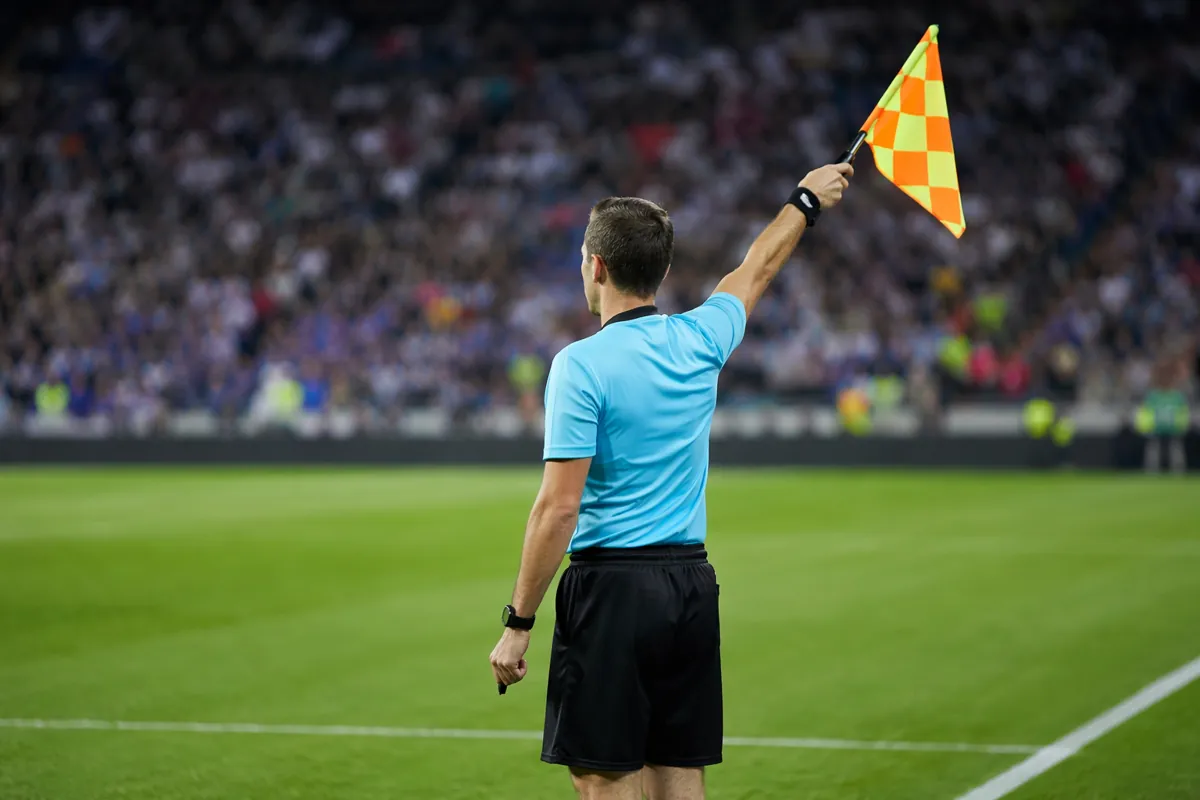 Editorial sports photograph of a soccer referee raising an offside flag on a green pitch,