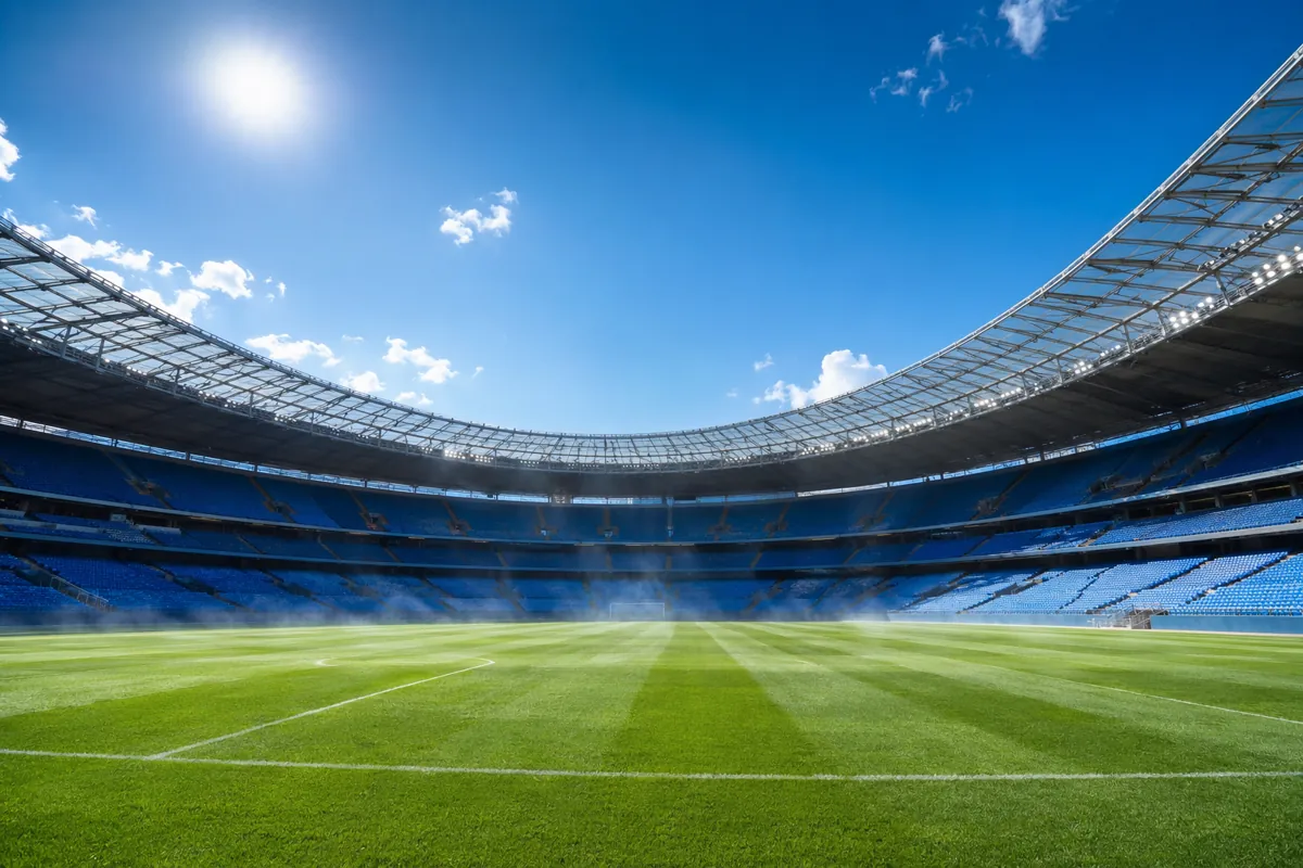 Wide shot of a sunlit professional football stadium during summer, bright blue sky, empty